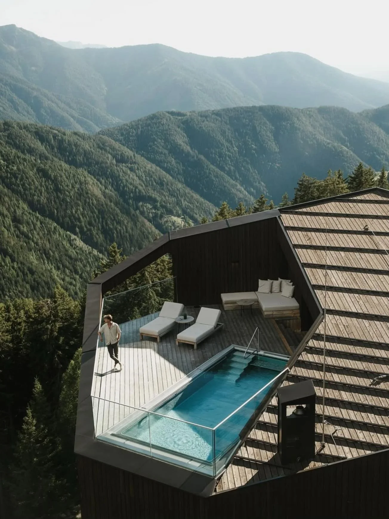 A modern house with a rooftop deck featuring a small swimming pool, lounge chairs, and a covered seating area with cushions, set against a backdrop of green mountains.