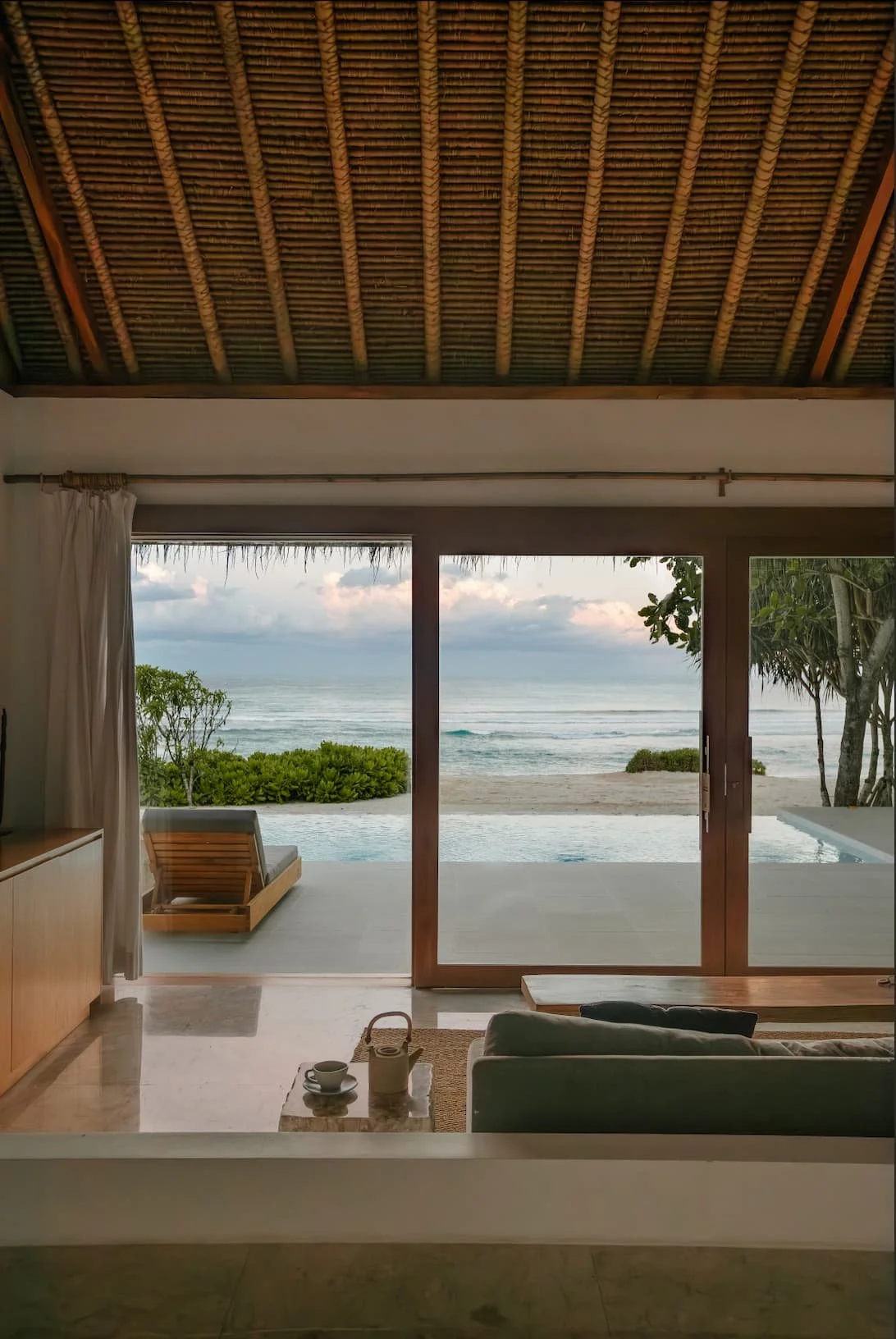 Indoor view of a room with large glass sliding doors opening to an outdoor patio, overlooking a beach with ocean waves, green bushes, and trees, with a thatched roof ceiling.