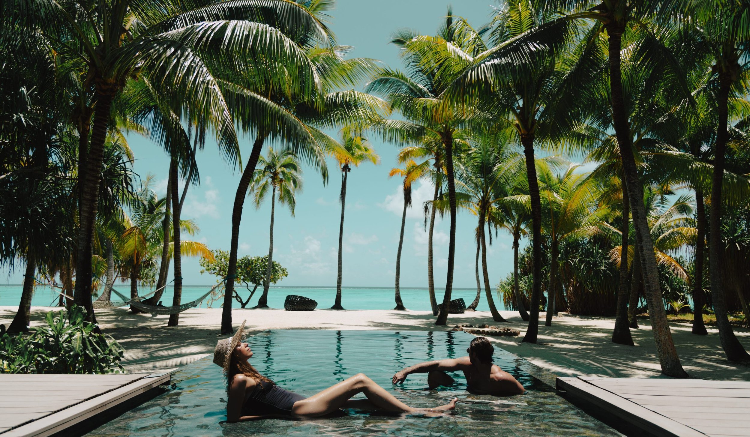 A couple relaxing in a tropical pool surrounded by palm trees with a view of the ocean in the background.