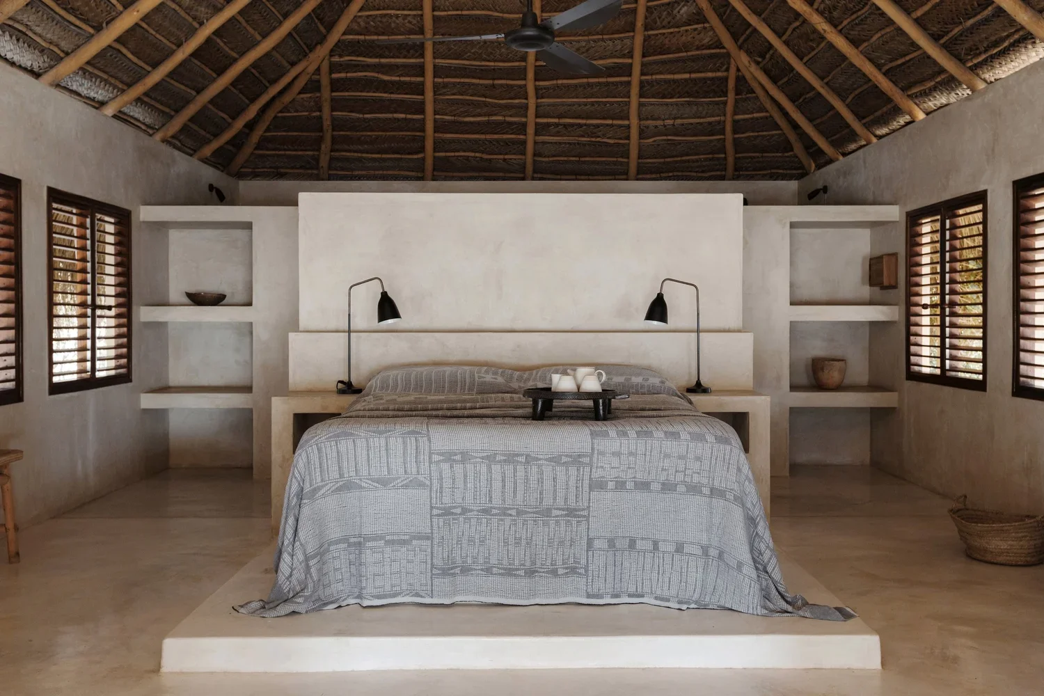 A bedroom with a high, thatched ceiling and neutral-toned walls. The bed has a patterned gray and white cover, with two black reading lamps on either side attached to a built-in headboard. A tray with white teacups is on the bed. There are built-in shelves with decorative bowls and boxes on each side of the bed. Four windows with wooden shutters are on the sides, and a ceiling fan hangs from the center of the ceiling.