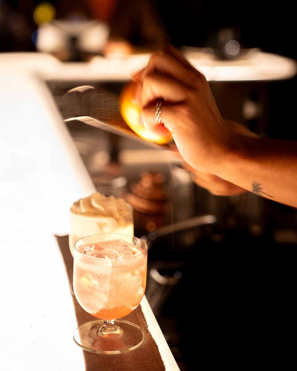 A bartender pouring a drink into a glass with ice, with a dessert and other drinks on the bar in the background.