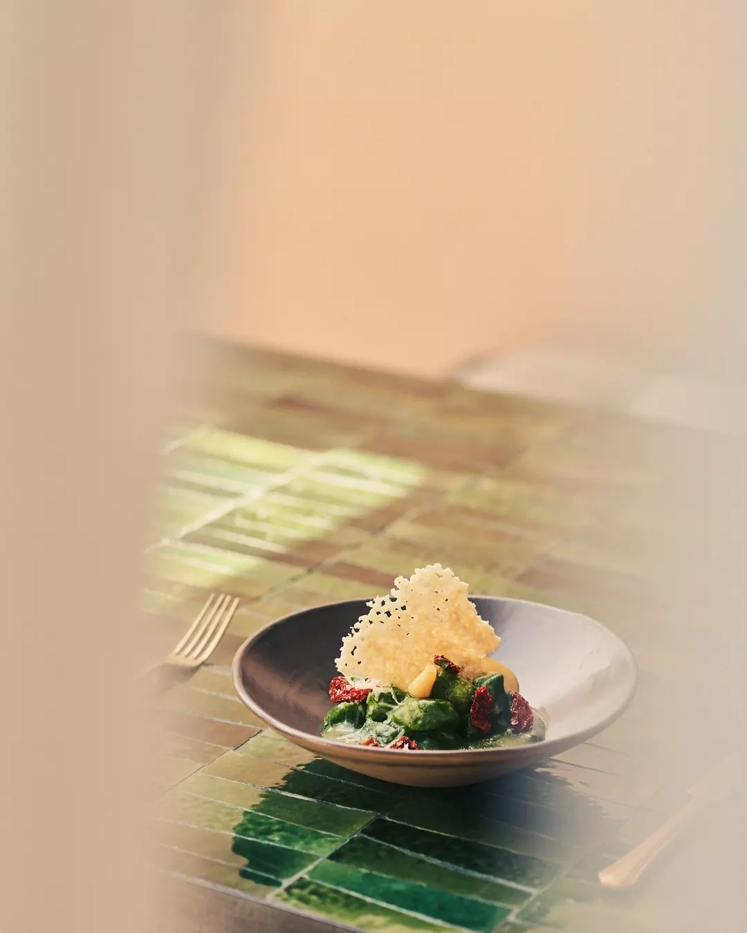 A black bowl containing a salad with leafy greens, dried cranberries, corn, and a crispy cheese tuile, placed on a green tile table with a fork beside it.