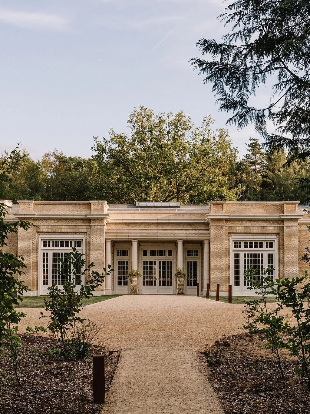 Brick building with large windows and a central entrance framed by columns, surrounded by greenery and trees.