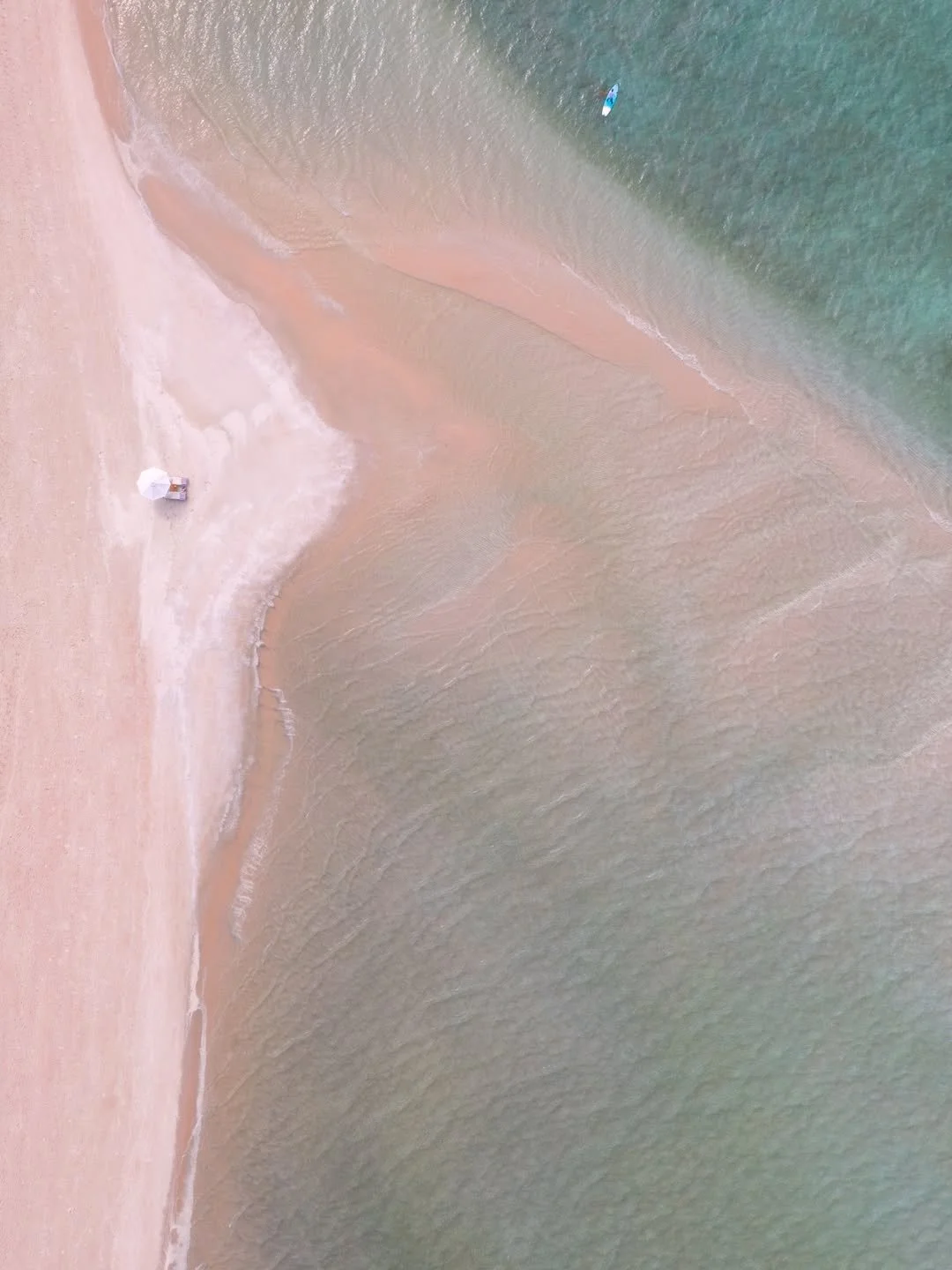 Aerial view of a beach with pinkish sand, small waves, a lone paddleboarder in the water, and a beach umbrella with a towel laid on the sand.