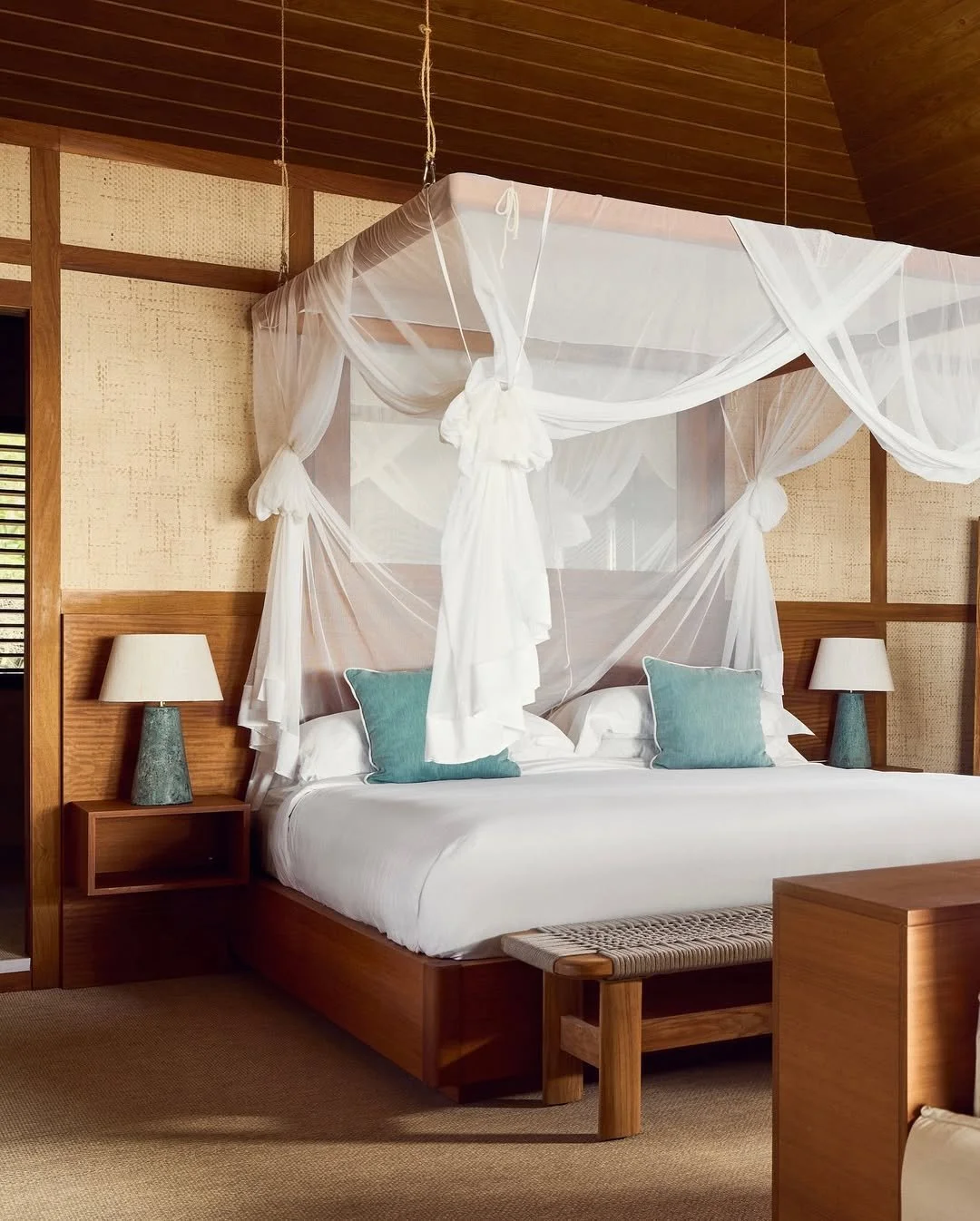A bedroom with a large bed featuring a white canopy, two turquoise pillows, and wooden headboard, flanked by two matching bedside tables with lamps, and a woven bench at the foot of the bed.
