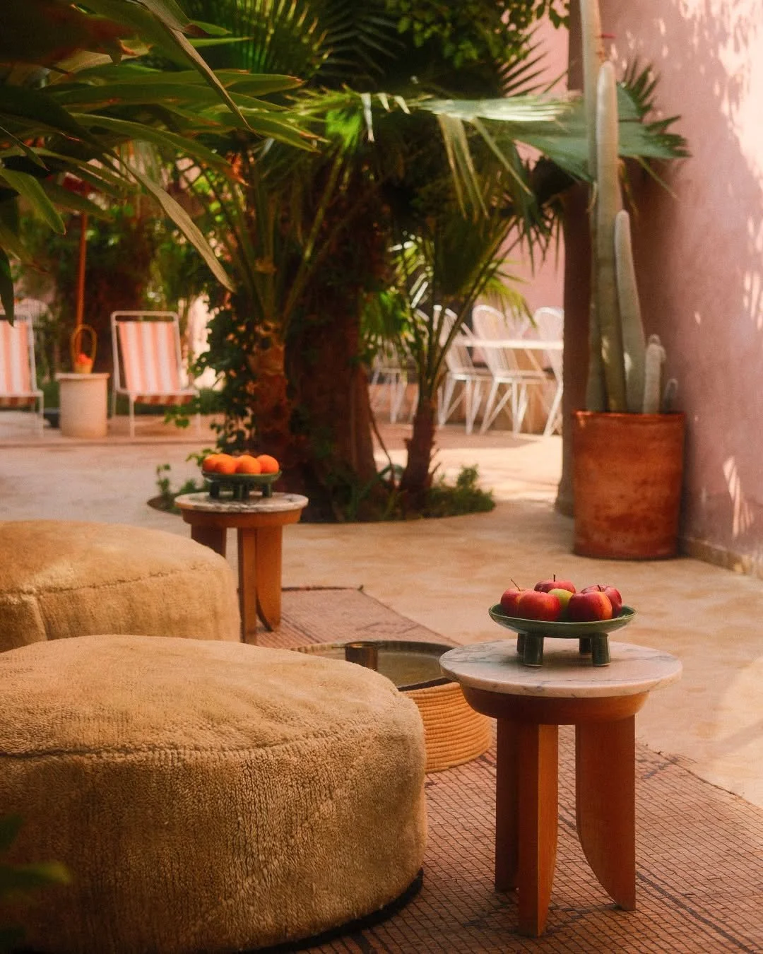 A cozy outdoor patio with two beige cushioned seats, two small round wooden tables with green plates of apples and peaches, lush green plants, including a palm and cactus, and white chairs in the background, bathed in warm natural light.