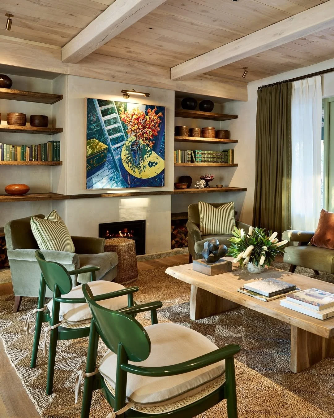 Living room with beige walls, wooden ceiling beams, green armchairs, a coffee table with books and decorative items, a fireplace, large window with green curtains, and built-in shelves with books and pottery, and a colorful painting on the wall.