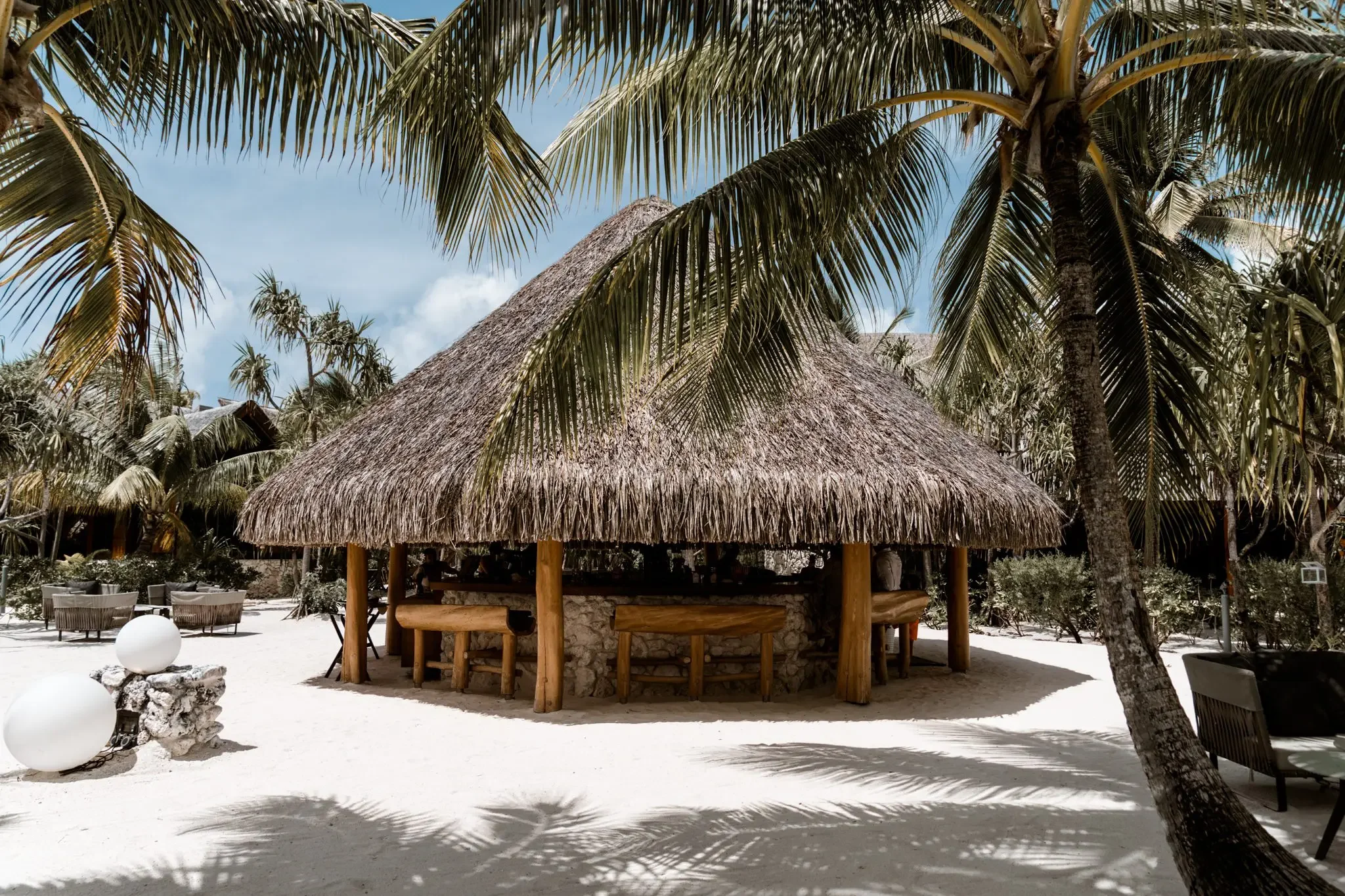 A tropical beach scene with a thatched roof hut, surrounded by palm trees and lounge chairs, under a bright blue sky.