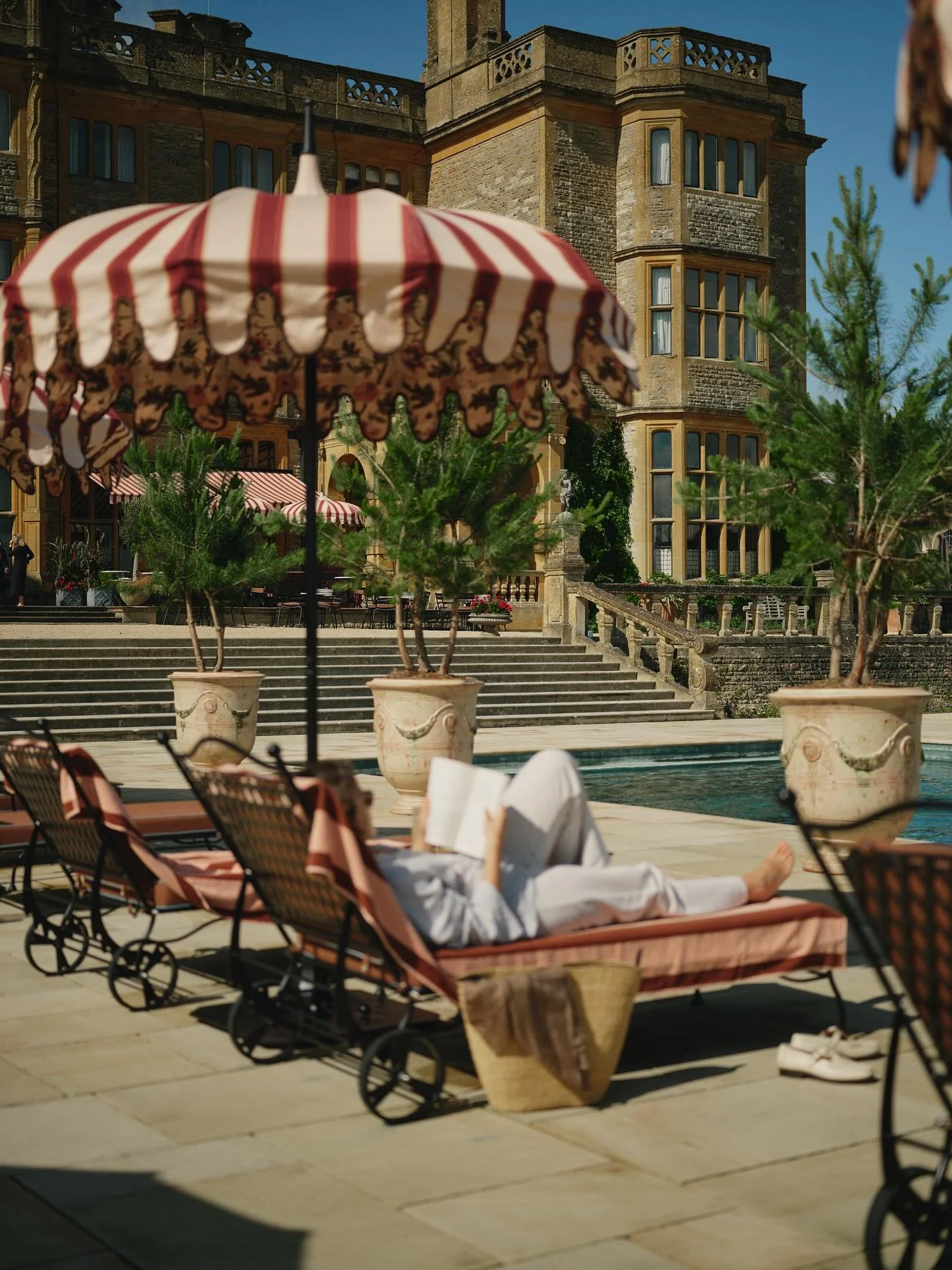 Person lying on a lounge chair by a swimming pool, with a red and white striped umbrella overhead. In the background, there is a large stone mansion with multiple windows, stairs leading up to it, and potted plants.