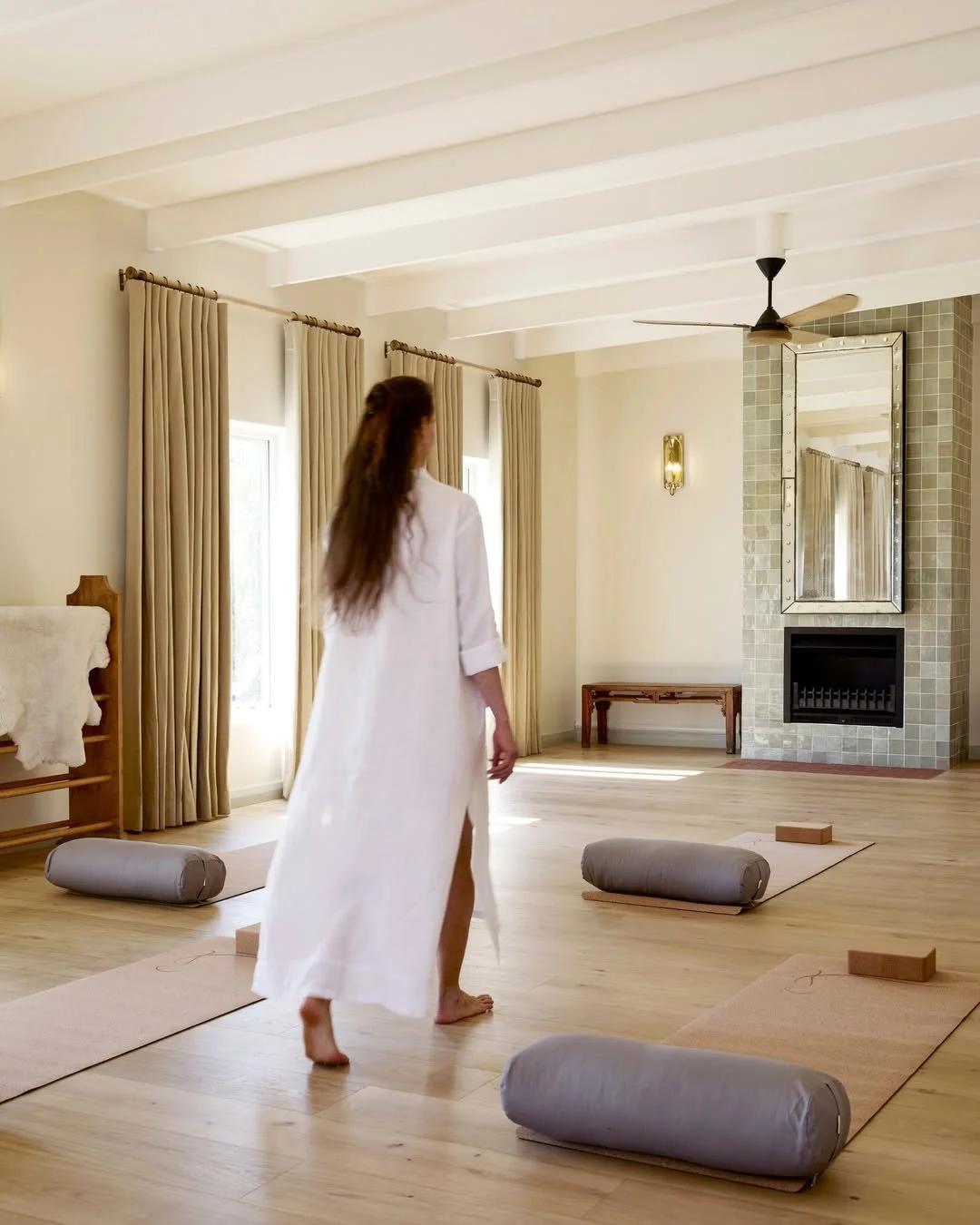 A woman walking barefoot in a spacious, bright room with yoga mats and bolsters, gold curtains, and a fireplace with a mirror above, decorated in a modern style.
