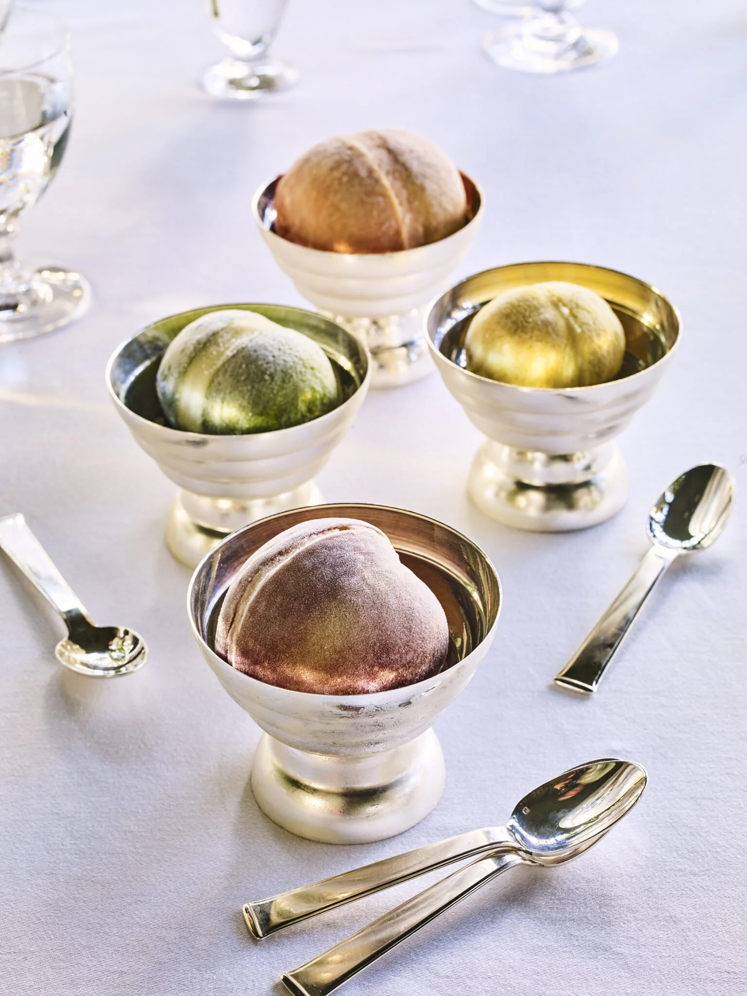 Three bowls of ice cream on a white tablecloth, with silver spoons nearby.