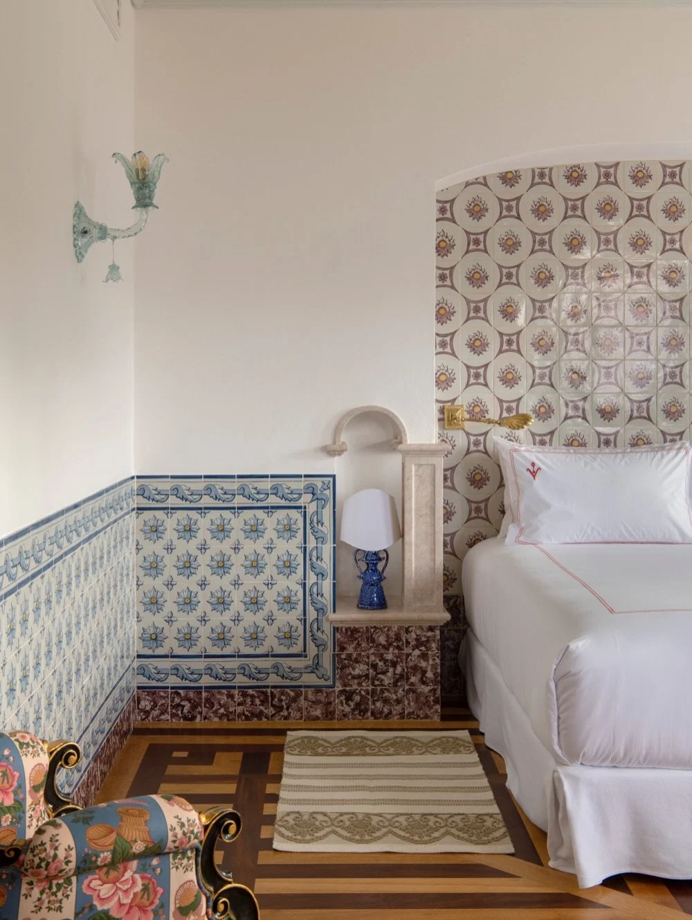 A bedroom corner with a white bed, a blue and white patterned wall with tiles, a small bedside lamp, a decorative armchair with floral upholstery, and a patterned rug on a wooden floor.