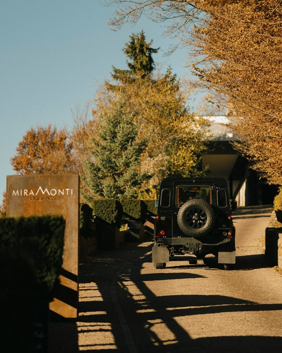 A black Land Rover Defender parked on a driveway surrounded by trees with fall foliage. There is a sign on the left with the text 'MIRA MONTI'.