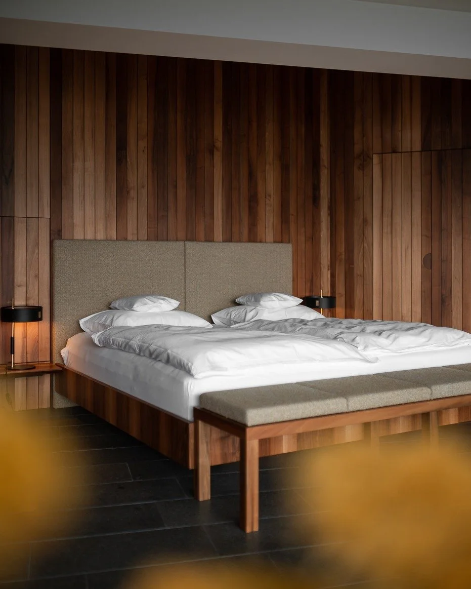Modern hotel bedroom with a large bed, beige headboard, and wooden wall paneling. Two black bedside lamps are on nightstands, and the bed has white linens and multiple pillows. A wooden bench sits at the foot of the bed.