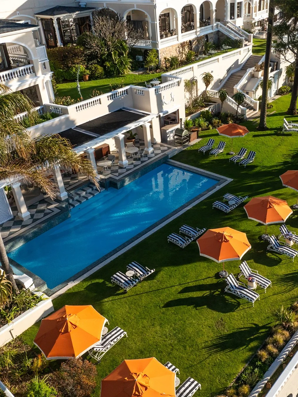 A luxurious hotel courtyard featuring a swimming pool surrounded by lounge chairs with orange umbrellas, lush green grass, palm trees, and a white multi-story building with balconies.