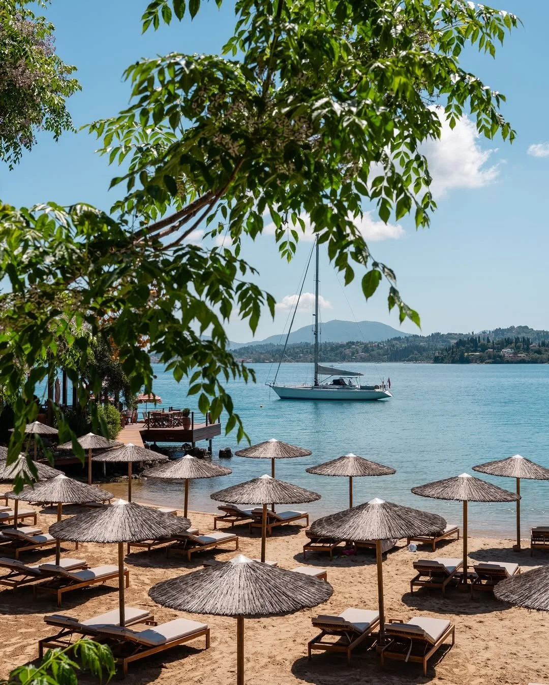 A beach with straw umbrellas and lounge chairs along the sandy shore, overlooking a calm lake with a sailboat, green trees, and mountains in the distance under a partly cloudy sky.