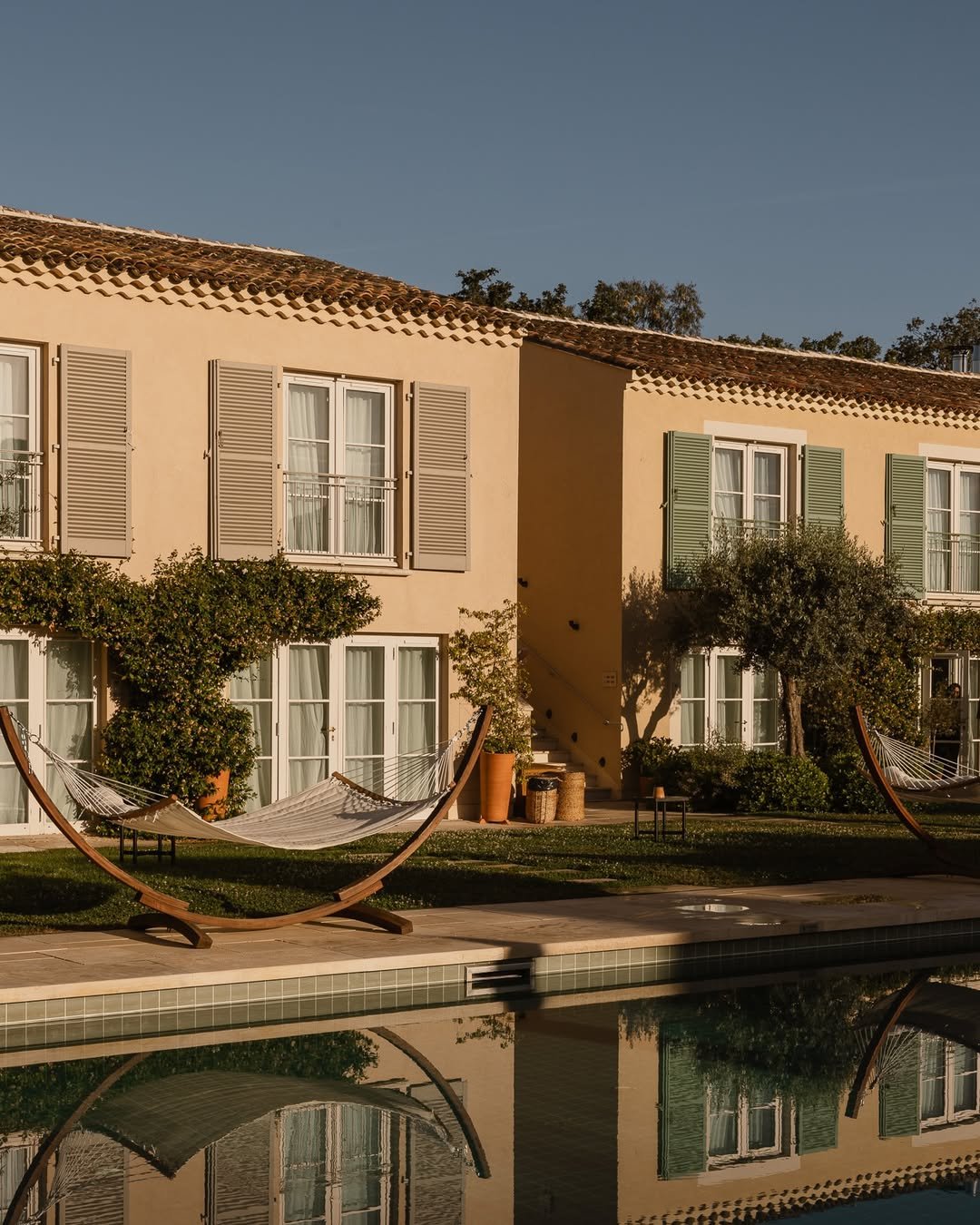 A poolside view of a beige apartment building with green shutters, hammock chairs, and trees in the yard during sunset.