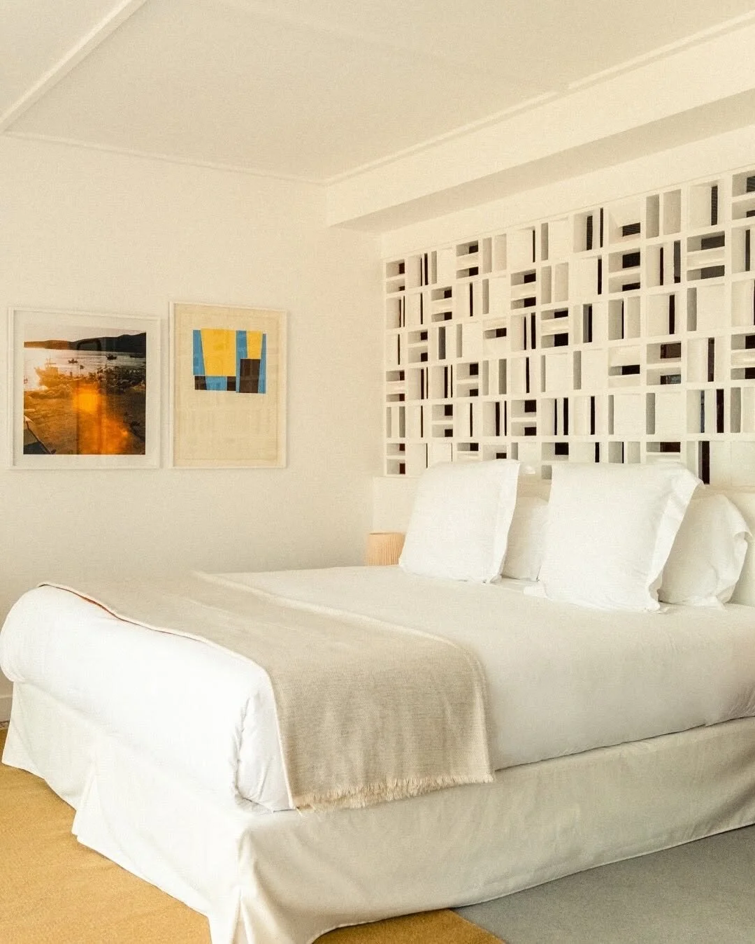 A bedroom with a white bed, beige blanket, and white pillows. There are two framed pictures on the wall, and a decorative white lattice headboard.