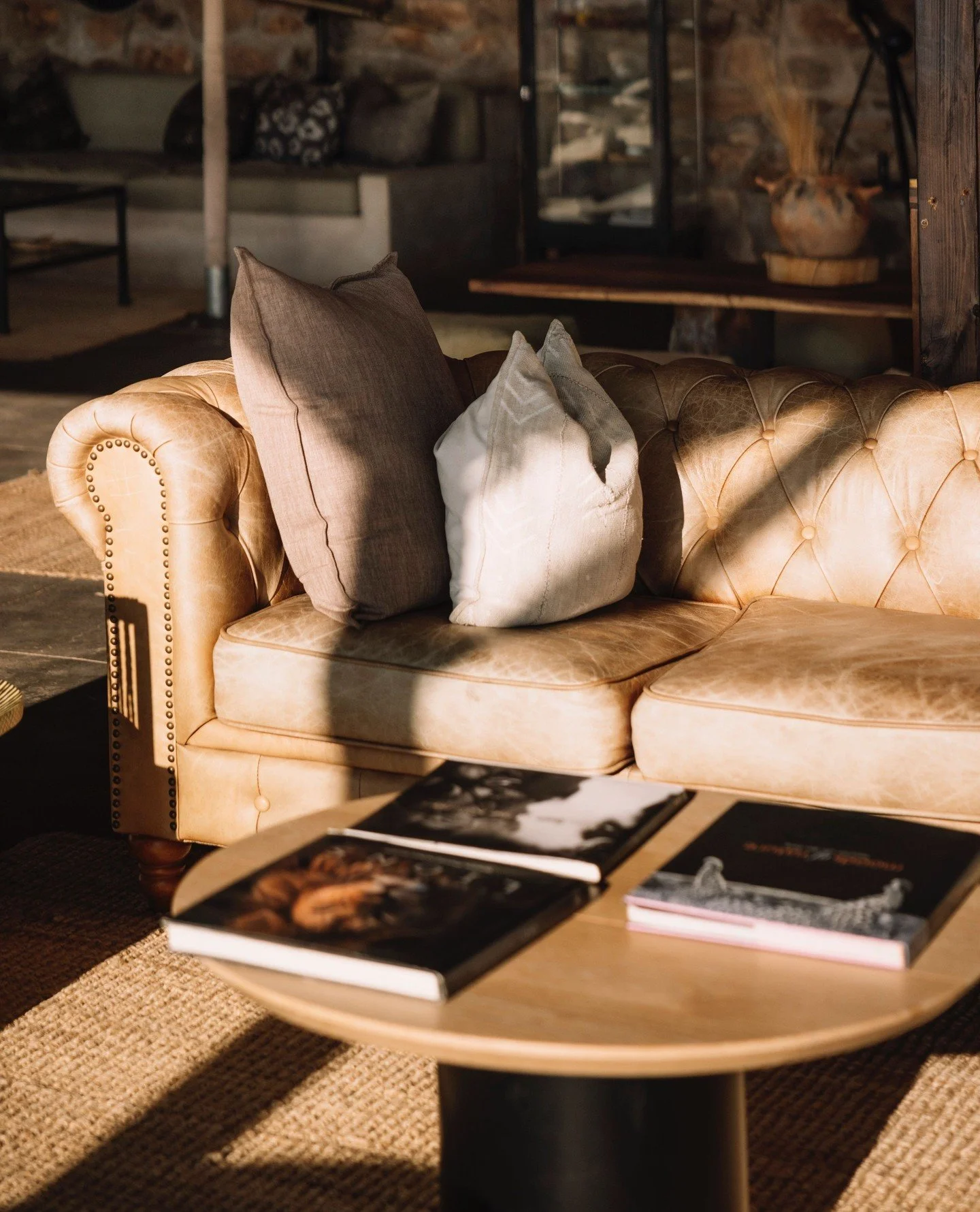 A beige leather tufted sofa with several throw pillows in gray, beige, and white. A wooden coffee table in front of the sofa has three books on it. The background includes a rustic wooden shelf with decorative items and a stone wall.