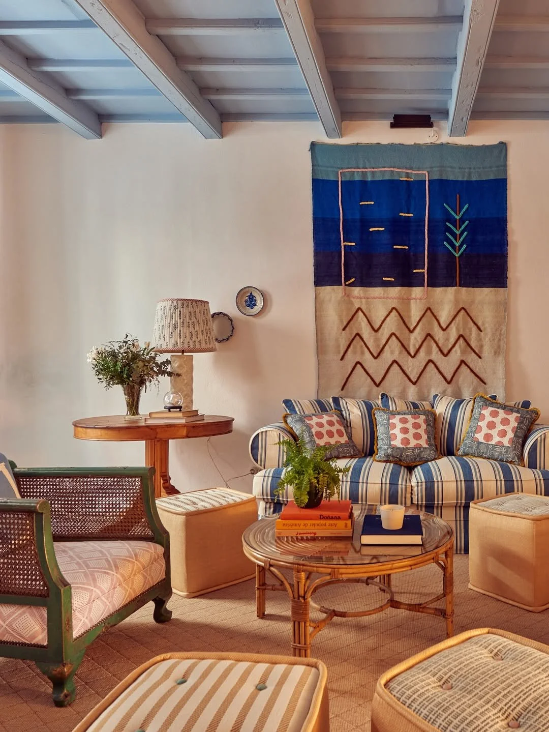 Cozy living room with a striped sofa, patterned cushions, a wooden coffee table with books, a potted fern, a side table with a vase and lamp, rattan furniture, and a colorful wall hanging.