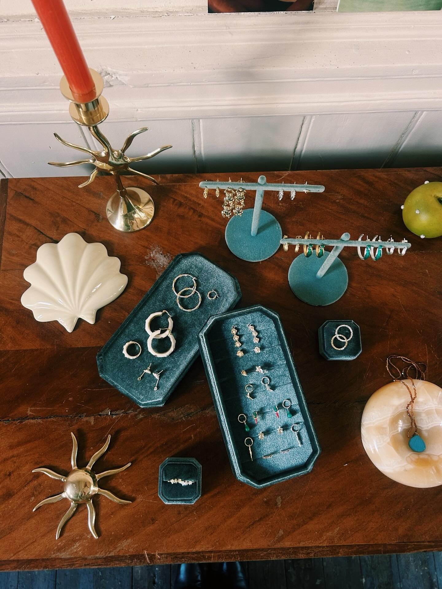 Jewelry display on a wooden table, featuring earrings and rings in various trays, a gold octopus-shaped candle holder, a green apple, and some seashell and starfish-shaped decorative objects.