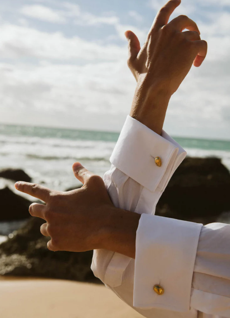 Close-up of a person's arms and hands with white shirt cuffs and gold cufflinks, making a gesture near the beach with rocks, sand, sea, and cloudy sky in the background.