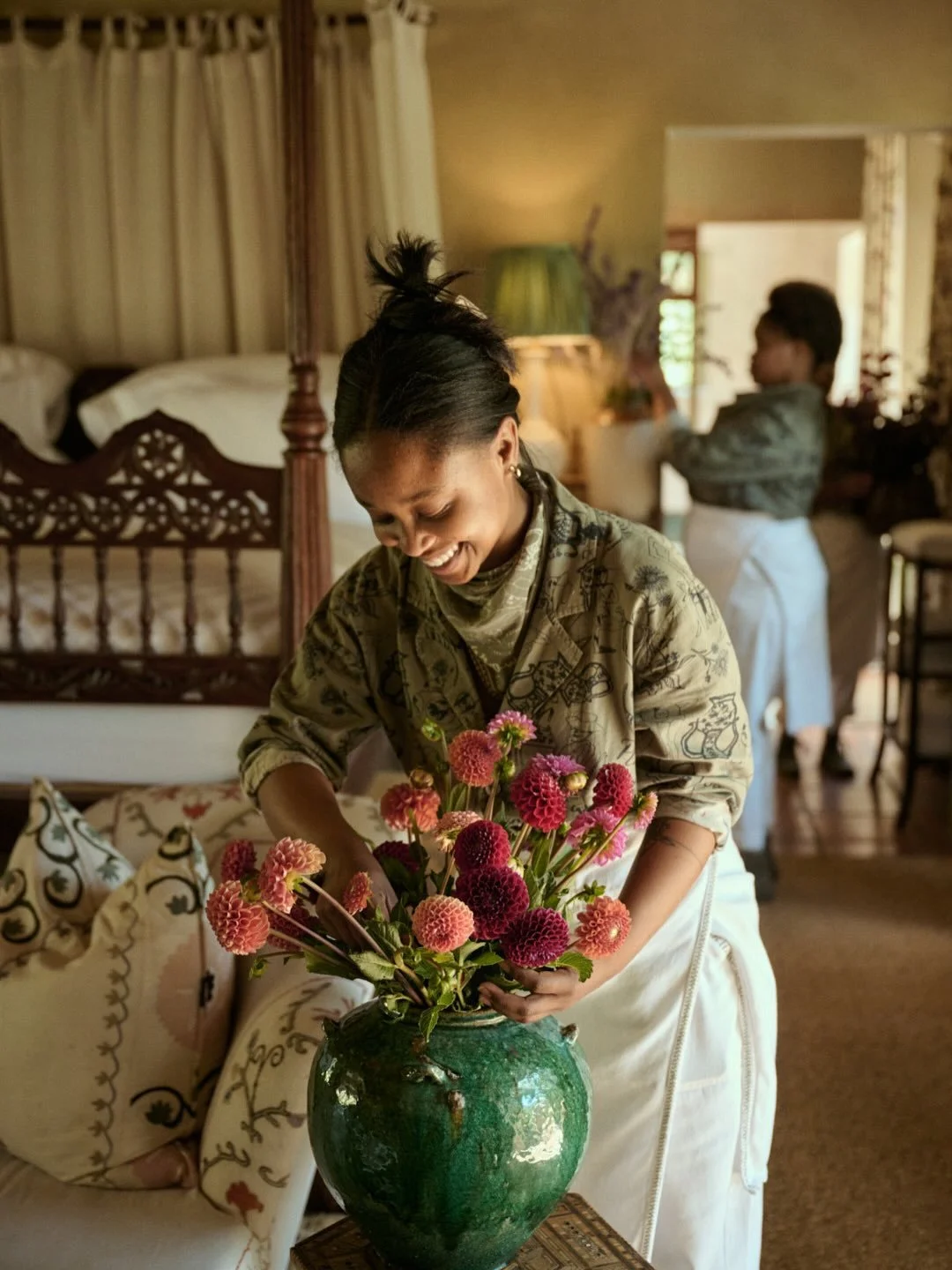 A woman arranging a bouquet of pink and red flowers in a green vase, smiling in a cozy home with a bed, curtains, and another person in the background.