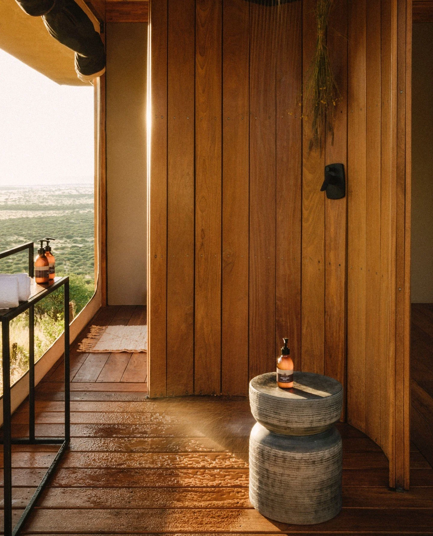 Indoor space with wooden walls and floor, a small round gray textured table with a pump soap bottle on top, and an open door leading to an outdoor view of greenery and hills.