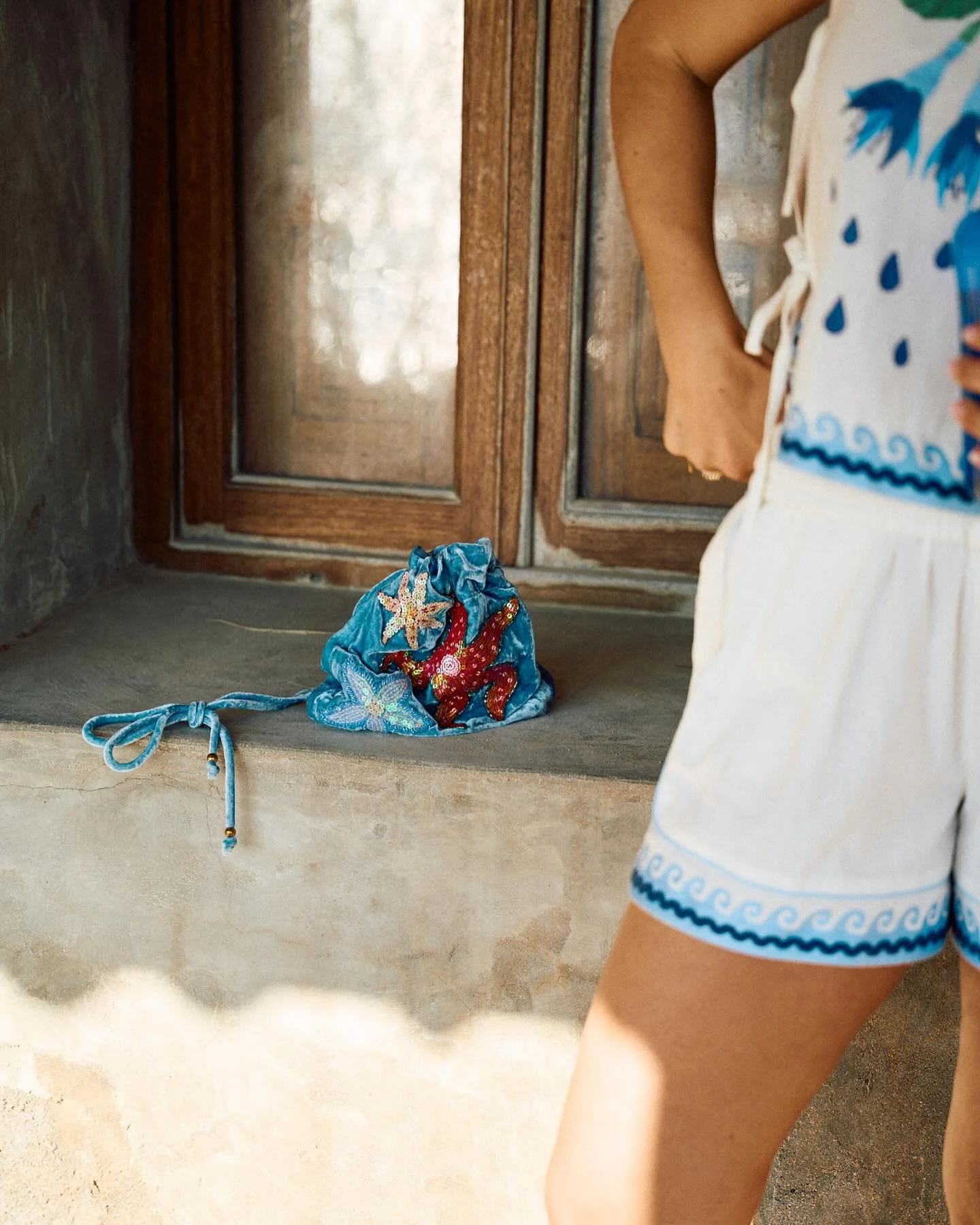 Part of a child's arm and torso in a white outfit with blue pattern, standing in front of a wooden door with a small blue drawstring bag with a starfish and octopus design resting on the concrete step.