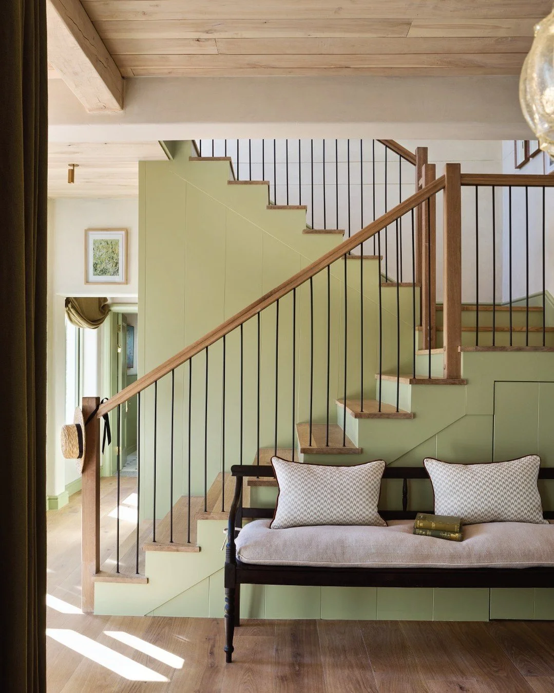 Interior of a house with a staircase, green wall, wooden railing, and a black bench with cushions and a book underneath.