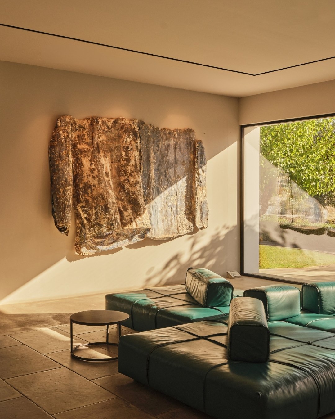 Modern living room with a green leather sectional sofa, a round coffee table, a large stone wall art piece, and a glass wall showing greenery outside, with sunlight casting shadows inside.