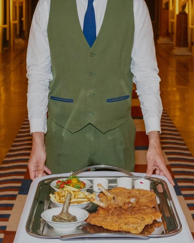 A waiter in a green vest, white shirt, and blue tie serving a tray with a large fried meat cutlet, a bowl of salad, and condiments in a restaurant hallway.