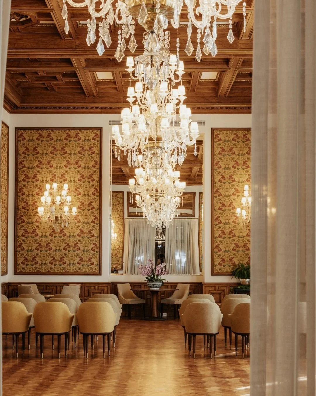 Elegant dining room with a high wooden ceiling, multiple crystal chandeliers, patterned wallpaper, beige chairs, and a large mirror reflecting the room's decor.