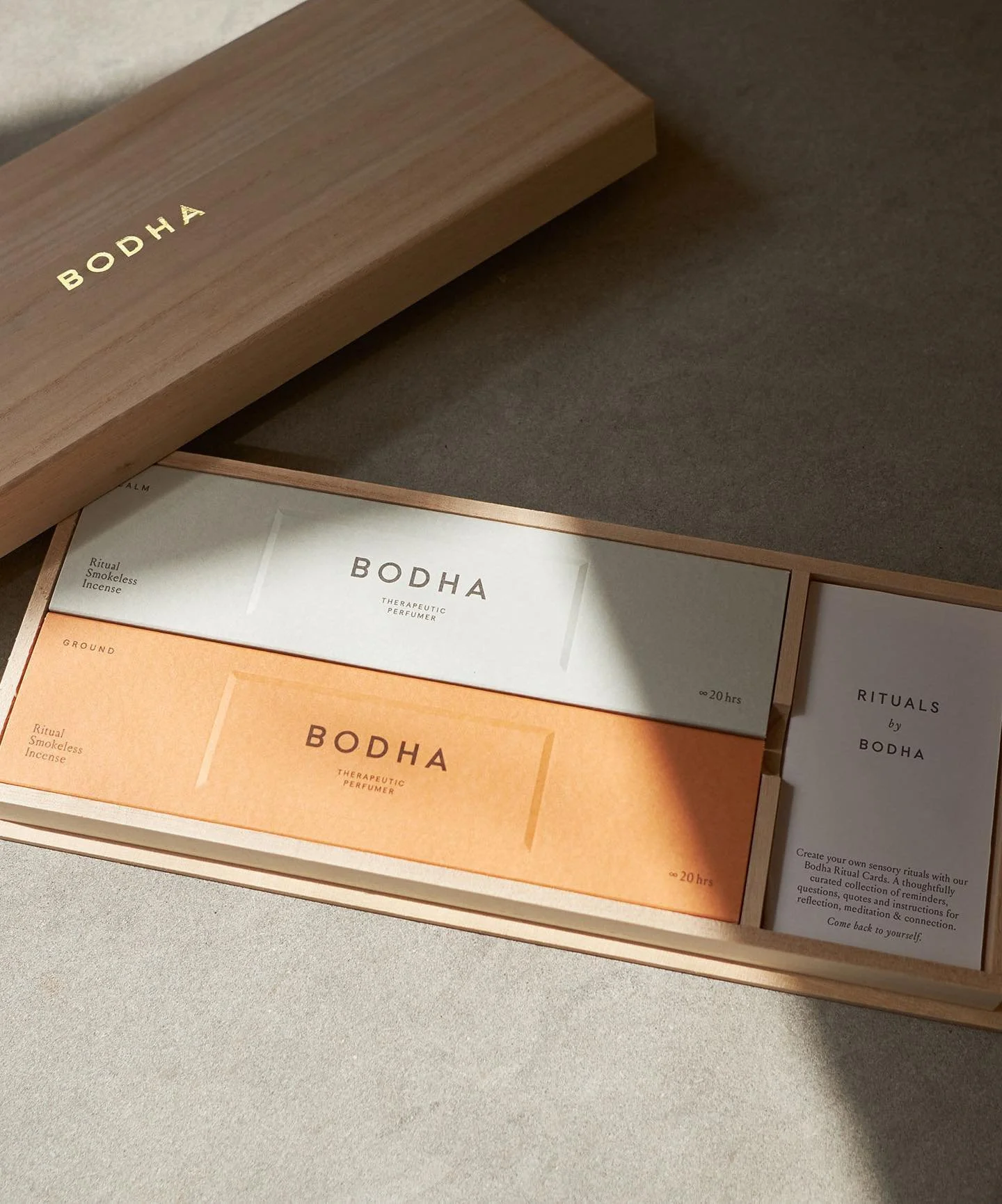 A wooden box with the brand name BODHA in gold letters, containing small boxes labeled Ritual Smokeless Incense, Therapeutic Perfumer, and other product descriptions, with a beige and white color scheme and minimalist design.