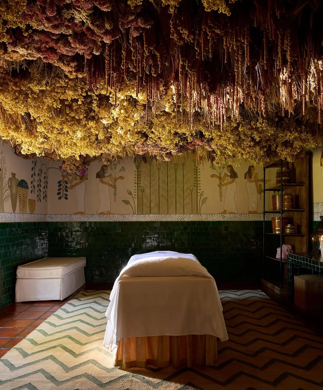 A room decorated with hanging dried flowers on the ceiling, a massage table draped with white cloth, green tile walls, and a mural of women and plants on the wall.