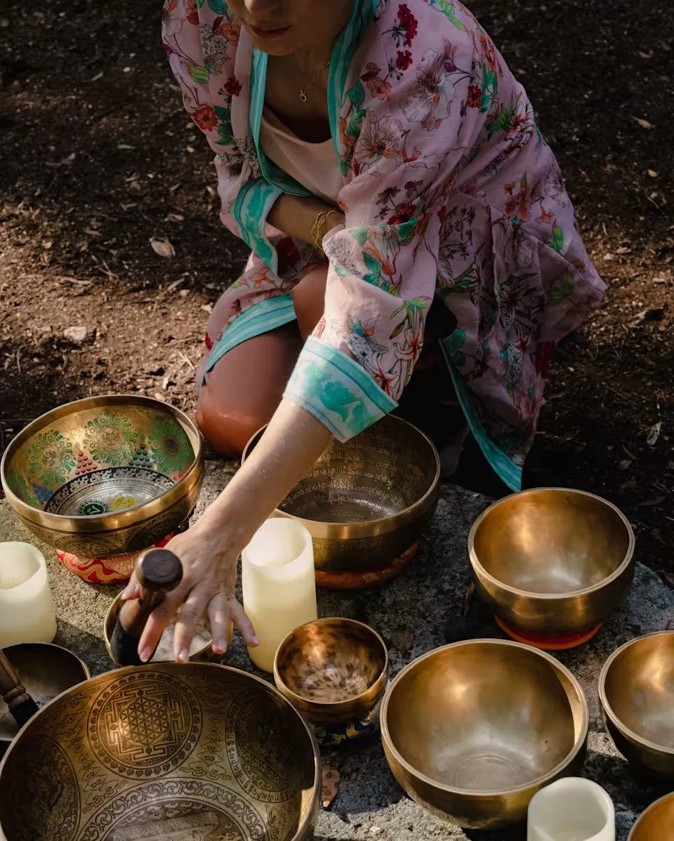 Person in a floral jacket playing singing bowls on the ground outdoors near candles and incense.