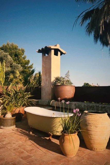 Outdoor patio with a white clawfoot bathtub, potted plants, and a chimney against a clear sky.