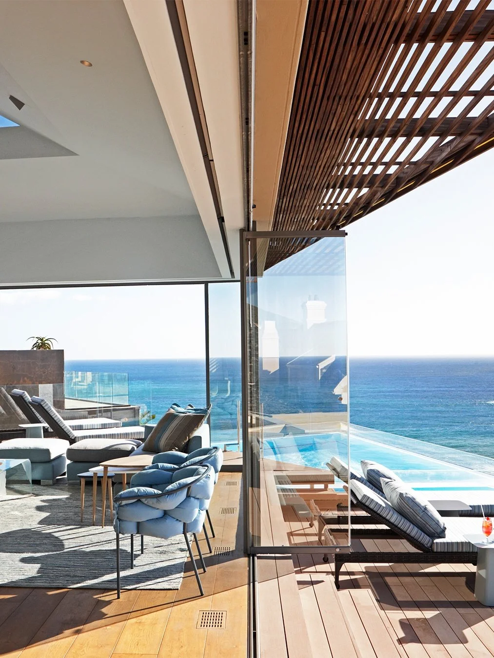 Modern living space with ocean view, outdoor seating area with chairs and loungers, glass railing, and wooden ceiling.