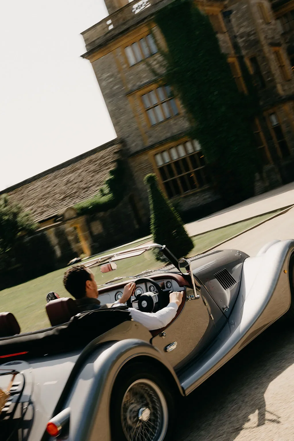 A man driving a vintage convertible car on a sunny day in front of a large stone building with ivy-covered walls and tall windows.