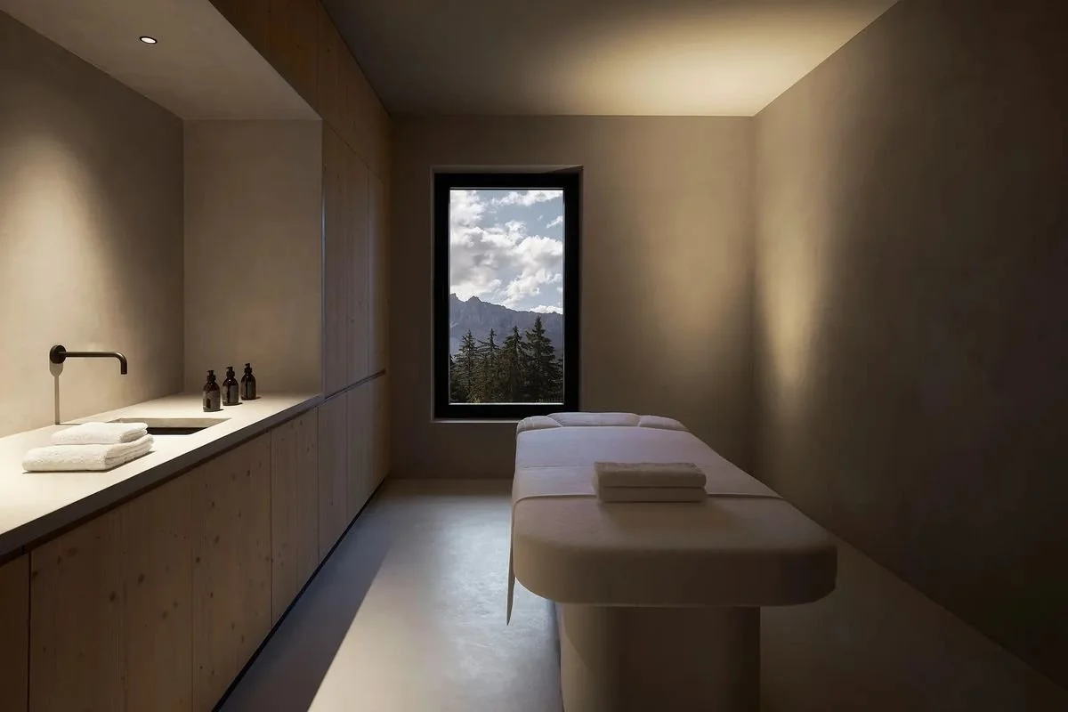 Minimalist massage therapy room with a massage table, window with mountain view, and cabinetry with bottles and towels.