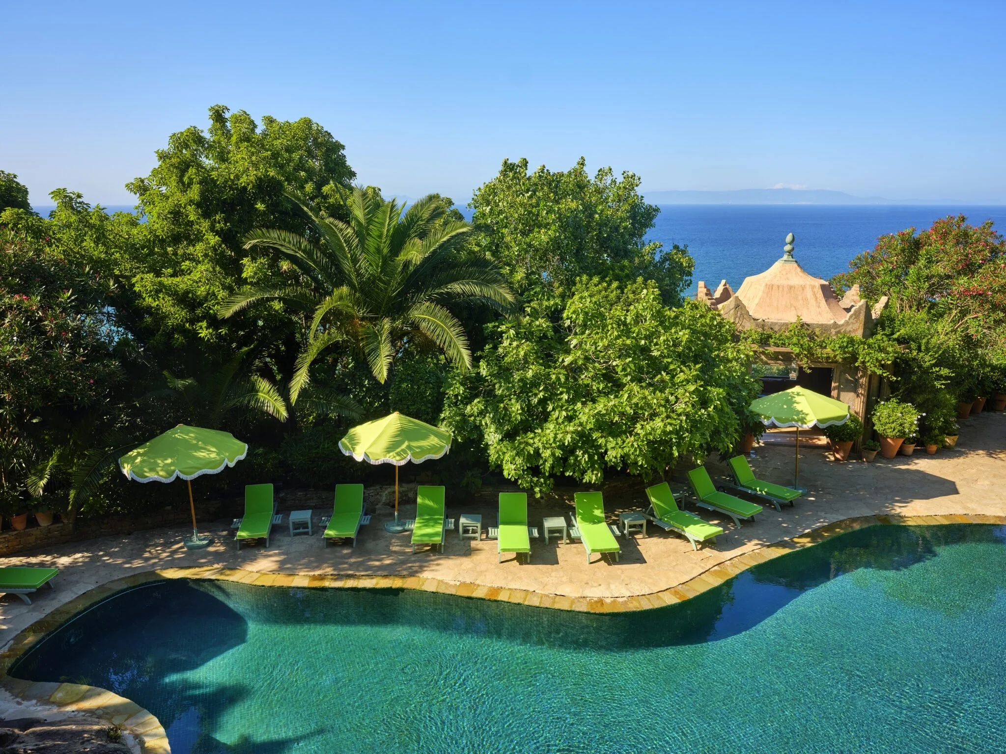 A tropical poolside area with a swimming pool, several green lounge chairs, umbrellas, lush green trees, and a view of the ocean in the background.