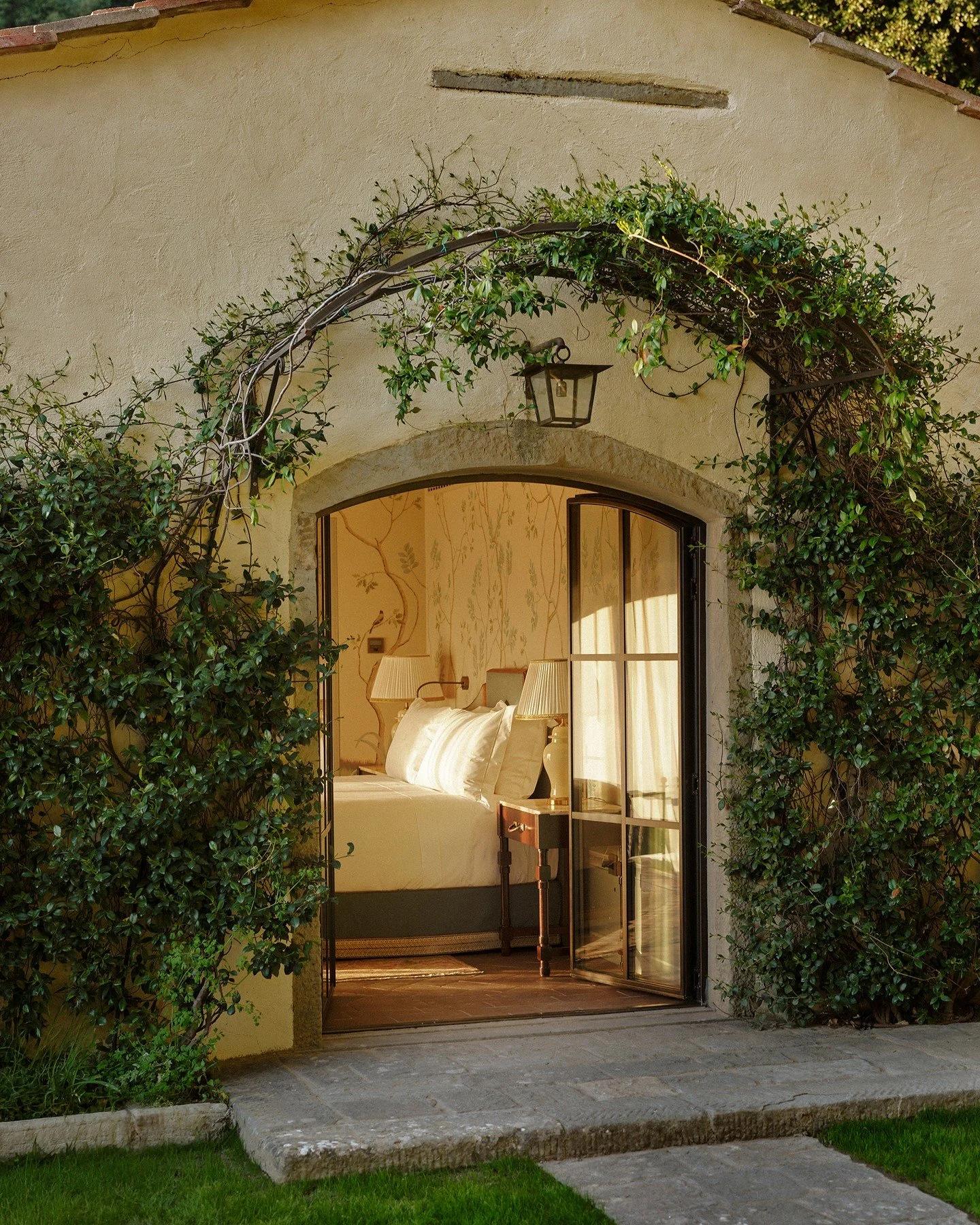 View through an open doorway showing a bedroom with a bed, lamp, and floral wallpaper, surrounded by green ivy on the exterior wall.