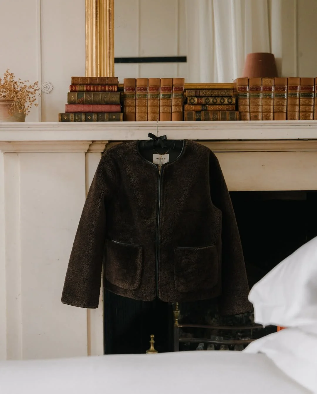 A brown fuzzy jacket hangs on a hook in front of a white fireplace, with a bookshelf filled with old books and a mirror above the shelf.