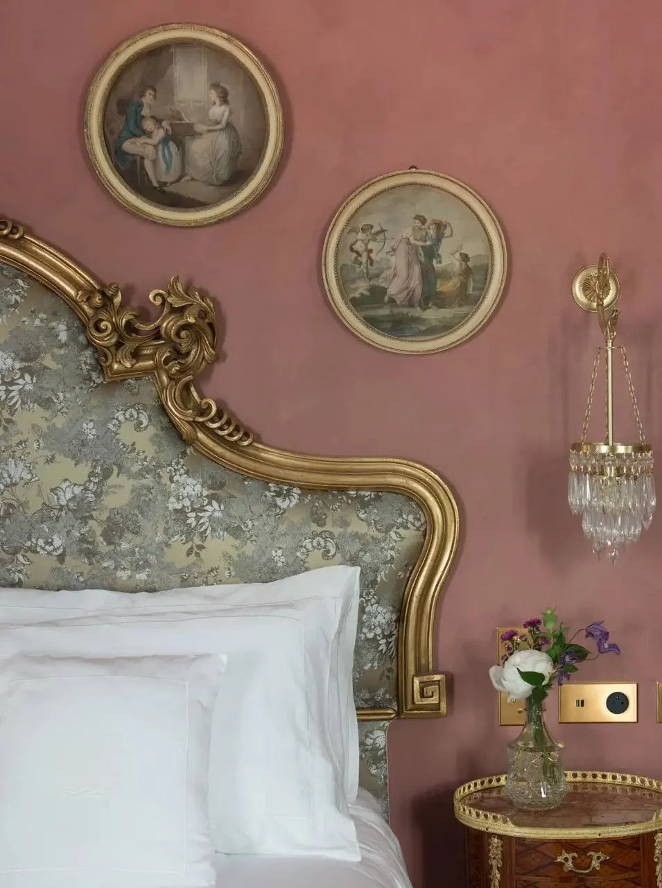 Luxurious bedroom with a floral upholstered headboard, gold trim, and pink walls decorated with vintage art prints. A side table holds a glass vase with flowers, and a crystal chandelier hangs beside wall outlets.