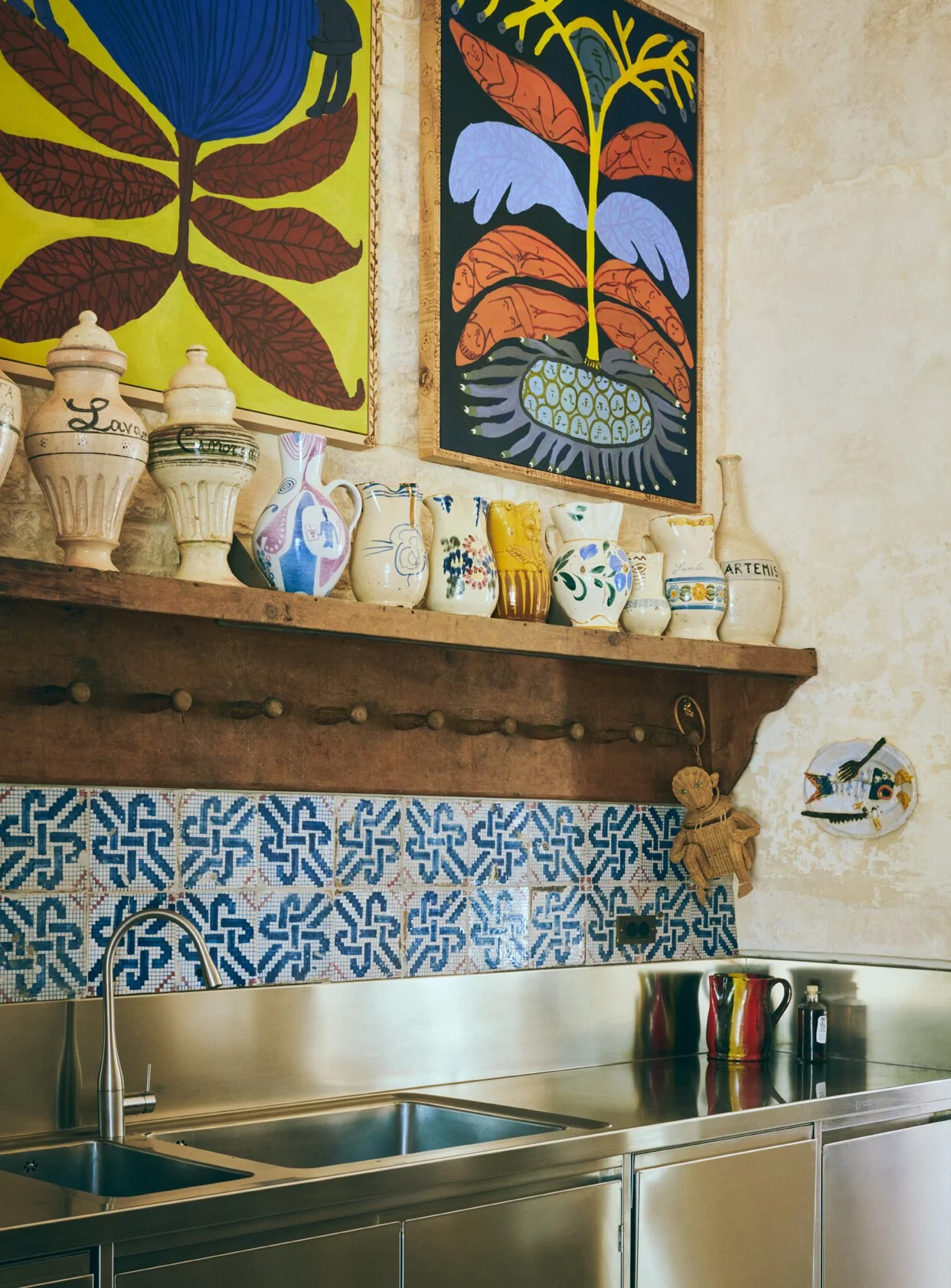 Decorative kitchen wall showcasing ceramic jars and colorful artwork, above a stainless steel sink and patterned tile backsplash.