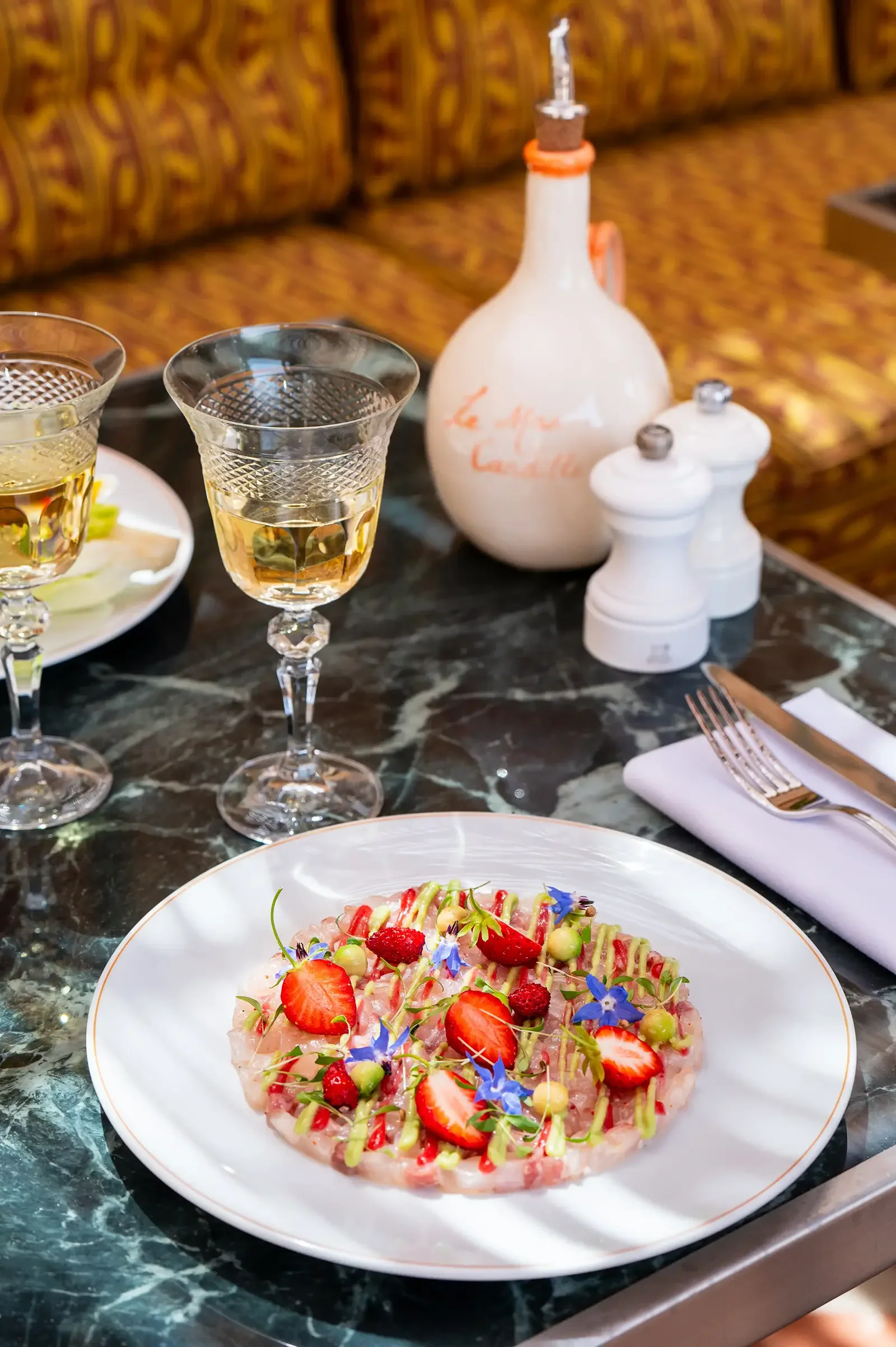 A fancy dinner table setup with two glasses of white wine, a decorative bottle, salt and pepper shakers, and a plated dish of seafood topped with strawberries, edible flowers, and drizzled sauce.
