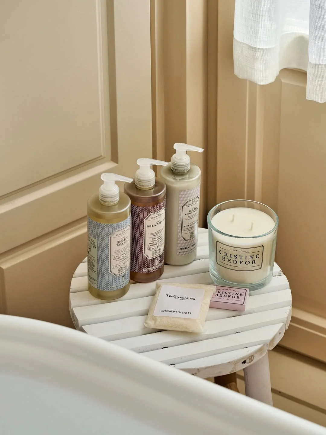 A small white wooden table with three bottles of shampoo, a candle labeled 'Cristine Bedford,' a bar of bath salts labeled 'TheGreenMood,' and a small box of soap beside the candle.
