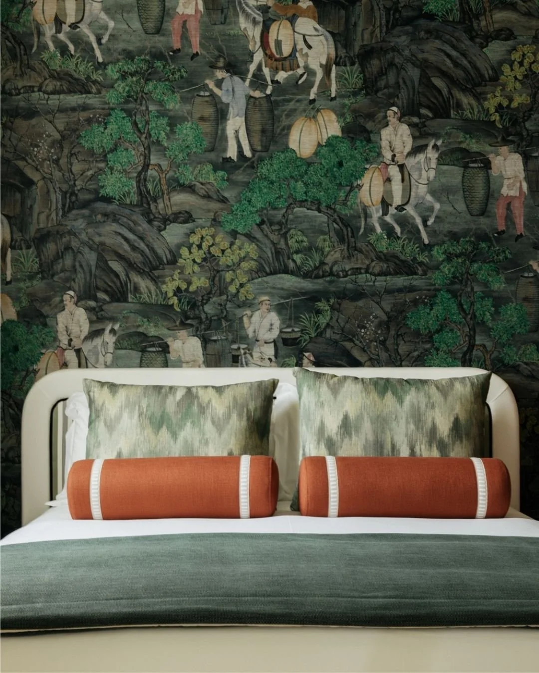 A bed with green and white pillows and orange cylindrical cushions, in front of a dark, intricate mural depicting traditional Asian figures and nature scenes with trees, rocks, and people carrying lanterns and goods.