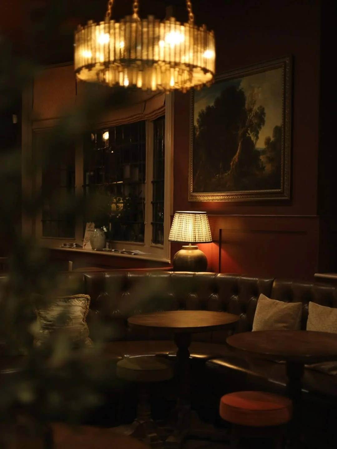 A dimly lit room with a circular chandelier hanging from the ceiling, soft lighting from a table lamp, dark leather sectional sofa, framed landscape painting on the wall, and large windows with blackout curtains.