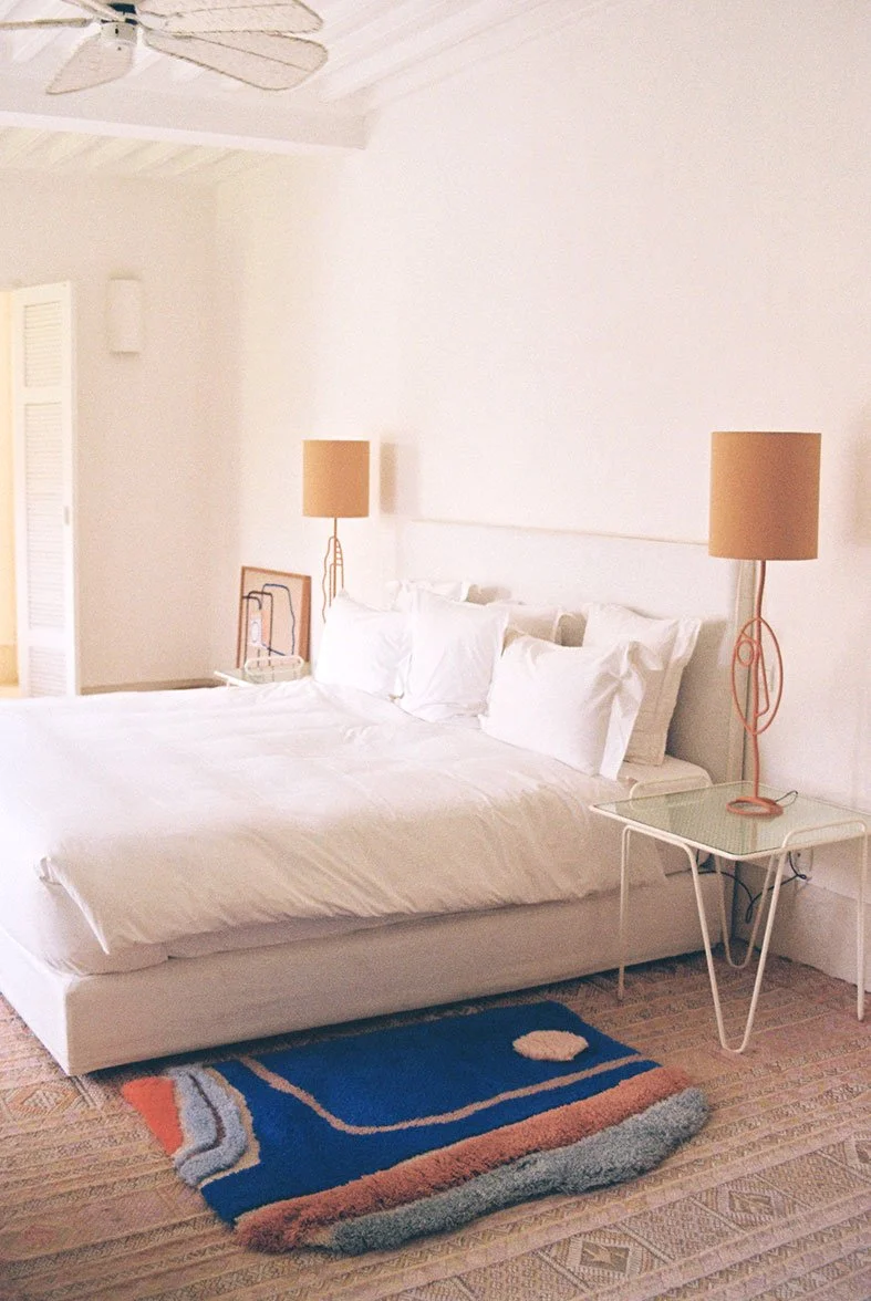 A neatly made white bed with multiple pillows, placed in a bright room with beige walls. Two matching beige lamps sit on white side tables on either side of the bed. A small colorful rug is on the floor at the foot of the bed, and a ceiling fan with wooden blades hangs from the ceiling.